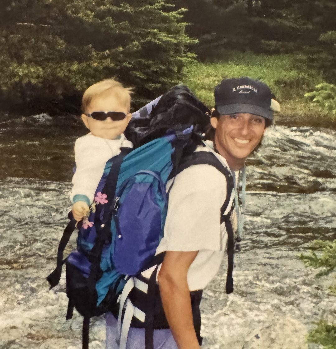 John riding in a hiking pack on his father's back beside a mountain river.